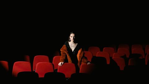 Woman in a black dress and a brown fur coat standing alone among rows of red seats in a dark, empty cinema, her hands resting on the seat backs in front of her