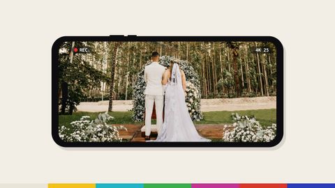 A phone held sideways recording an outdoor wedding ceremony, the couple seen from behind at a flower arch at the end of a wooden aisle